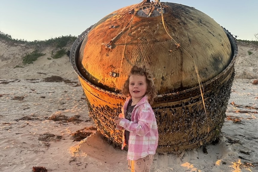 A little girl with curly brown hair and a pink jacket stands next to a big cylinder on a beach