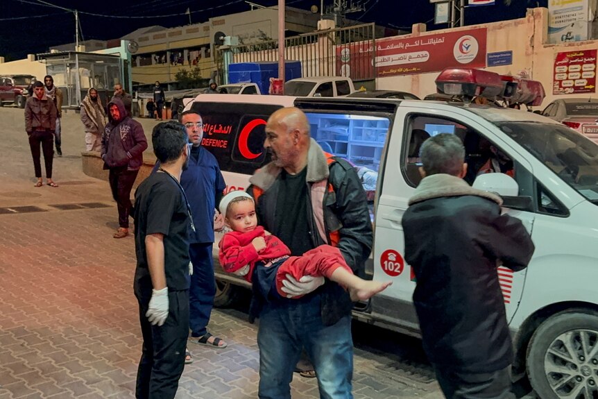 A child is carried from an ambulance by a man as hospital staff stand nearby 