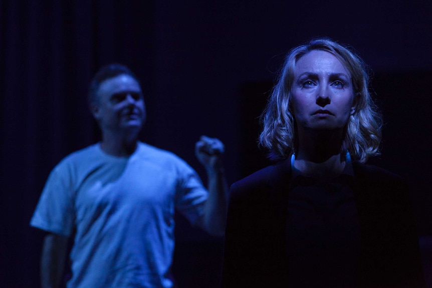 The actors stand, looking up sadly, bathed in blue light and shadow.