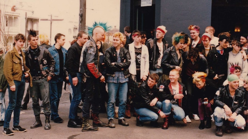 About 20 punks, some with leather jackets and colourful mohawks, stand in front of a pub. They are smiling.