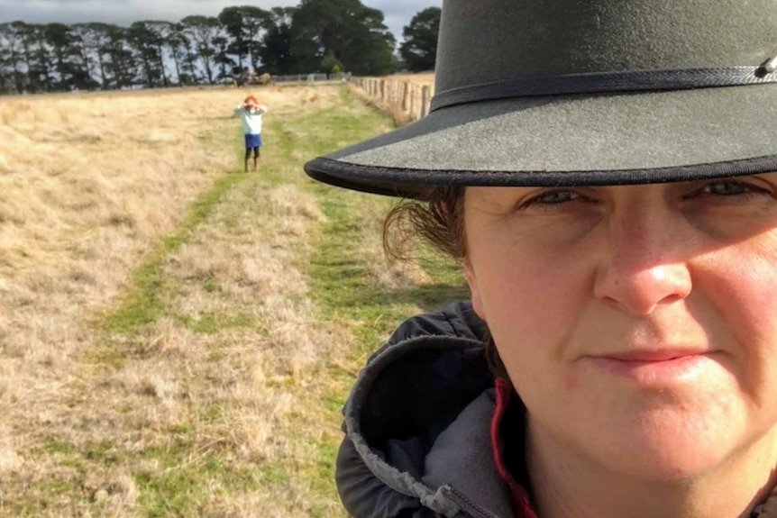A woman wearing an Akubra takes a selfie close to the camera with her farm in the background.