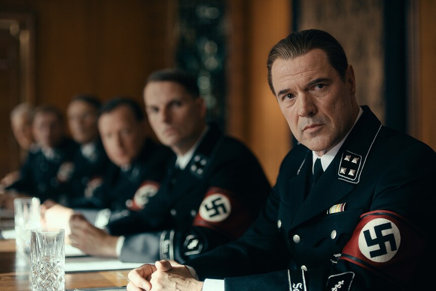 Colour still of Sebastian Koch sitting at table with 5 other men wearing Nazi uniforms in 2018 film Never Look Away.