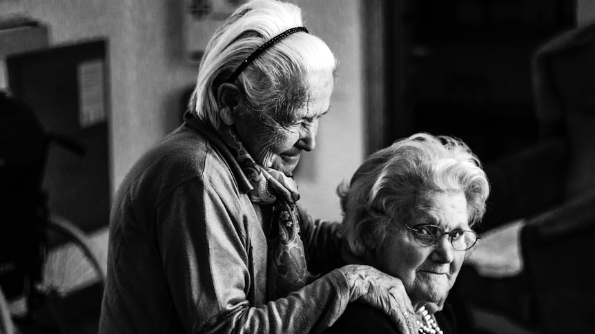 B&W photo of two older women, one of who is resting her hands on the shoulders of the other.
