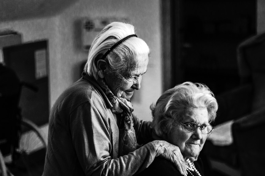 B&W photo of two older women, one of who is resting her hands on the shoulders of the other.