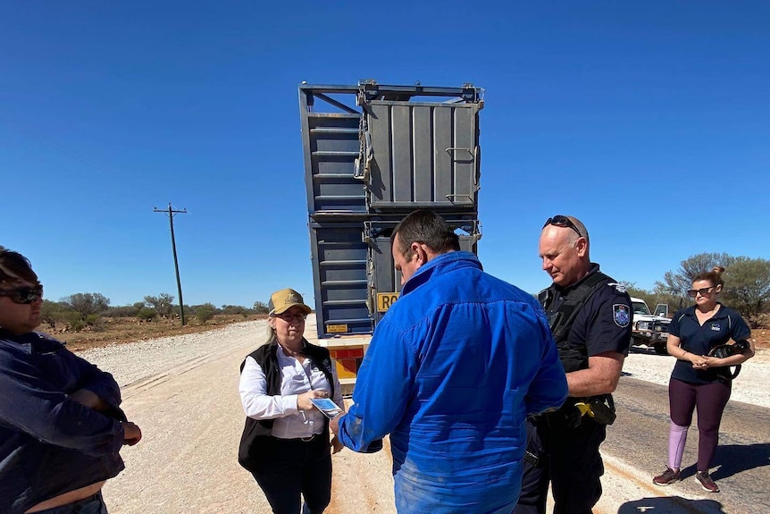 A lady in a trucker hat hands a road train driver a brochure in the outback