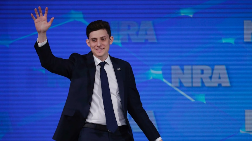 Kyle Kashuv waves as he stands in front of a screen with NRA logos.