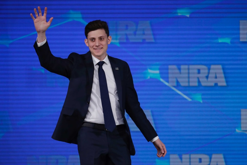 Kyle Kashuv waves as he stands in front of a screen with NRA logos.