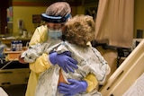 A healthcare worker wearing a mask and protective screen and scrubs hugs a woman in a hospital gown sitting on a bed.