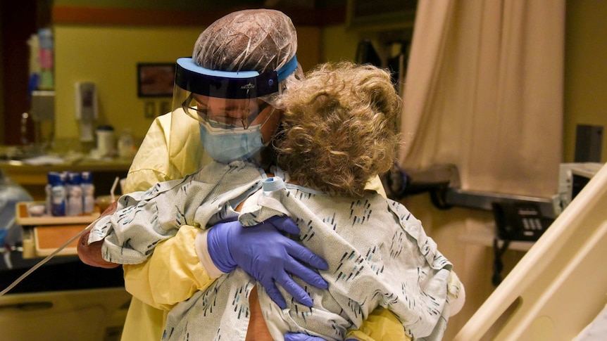 A healthcare worker wearing a mask and protective screen and scrubs hugs a woman in a hospital gown sitting on a bed.