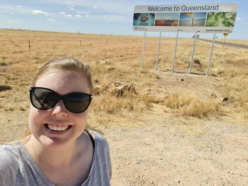 A woman with sunglasses smiles in front of a sign that says welcome to Queensland.