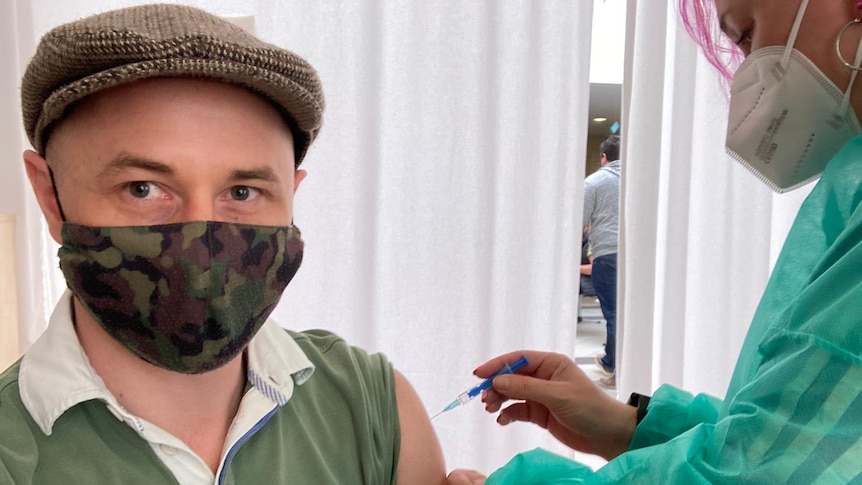 A man wearing a hat and mask receives a vaccine from a nurse with pink hair.
