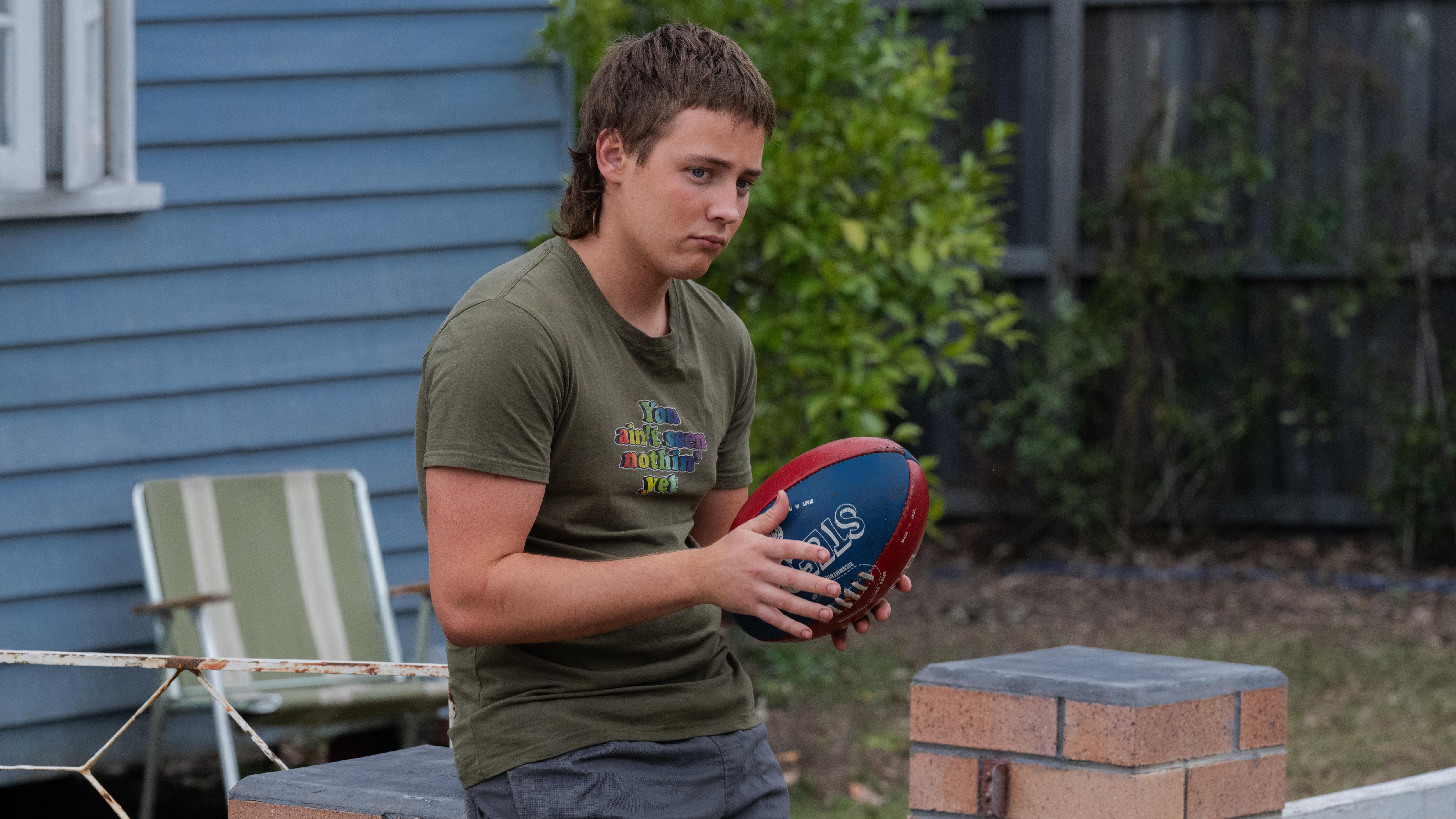 Gus holds a football in his hands and looks into the distance while leaning on a brick fence in front of his house.