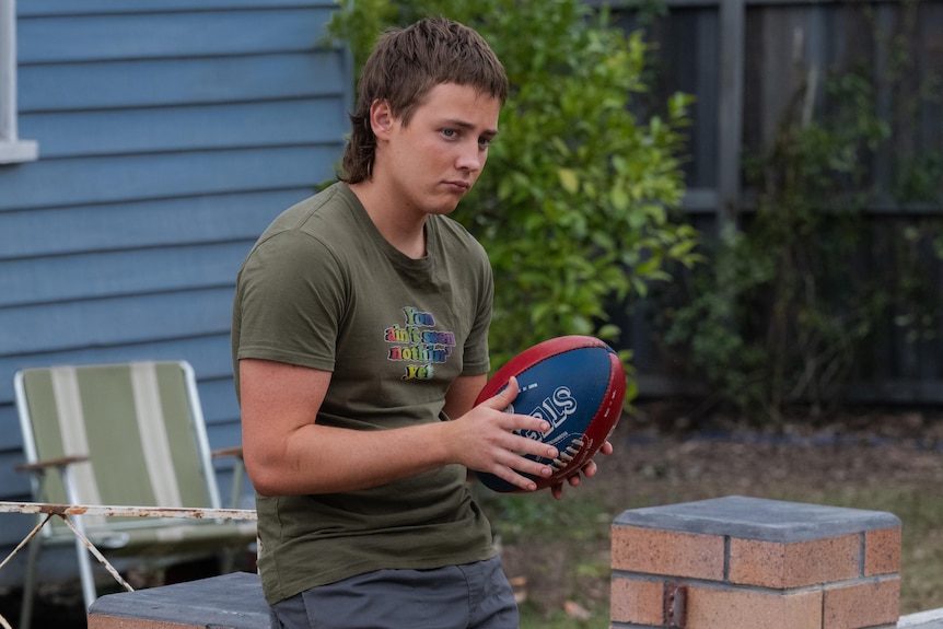 Gus holds a football in his hands and looks into the distance while leaning on a brick fence in front of his house.