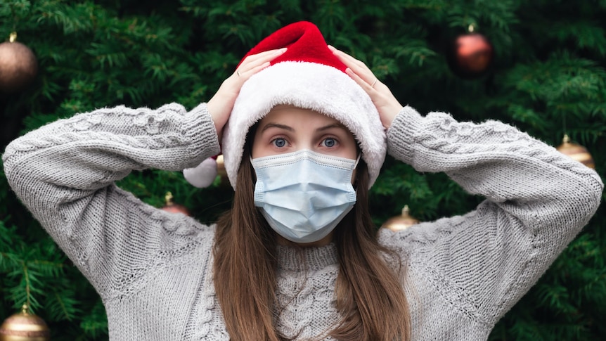 Close up Portrait of woman wearing a santa claus hat and medical mask against the background of a Christmas tree.