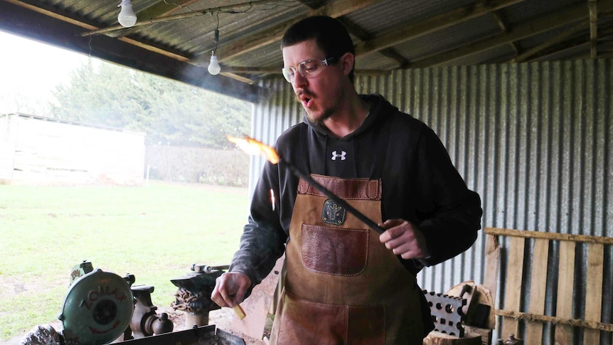 a blacksmith holds a steel rod in one hand and a chunk of beeswax in the other