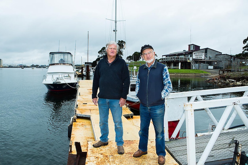 Two older men stand on a jetty near a jetty next to a river, with a yacht club building in the background.