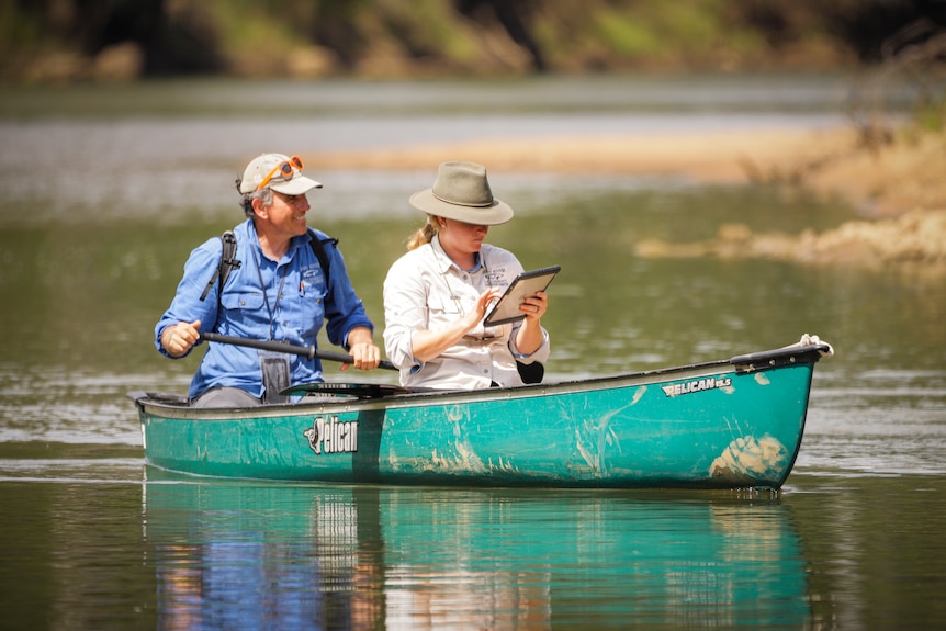 Teal canoe on a river with a man in the back and a woman in front, looking at a tablet. 