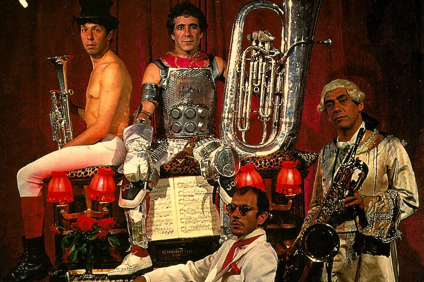 Four men dressed in different stage costumes, including a suit, and armour, sit together holding instruments.