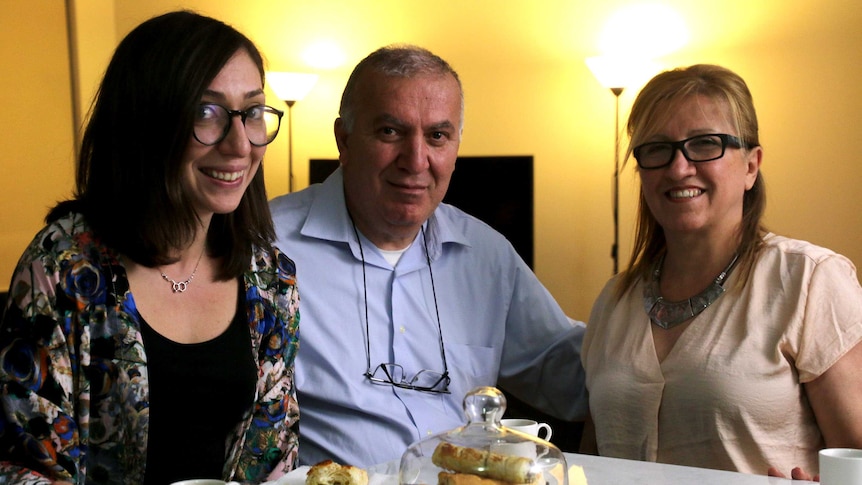 Talar sits with her father and mother at a table with pastries and coffee in the foreground.