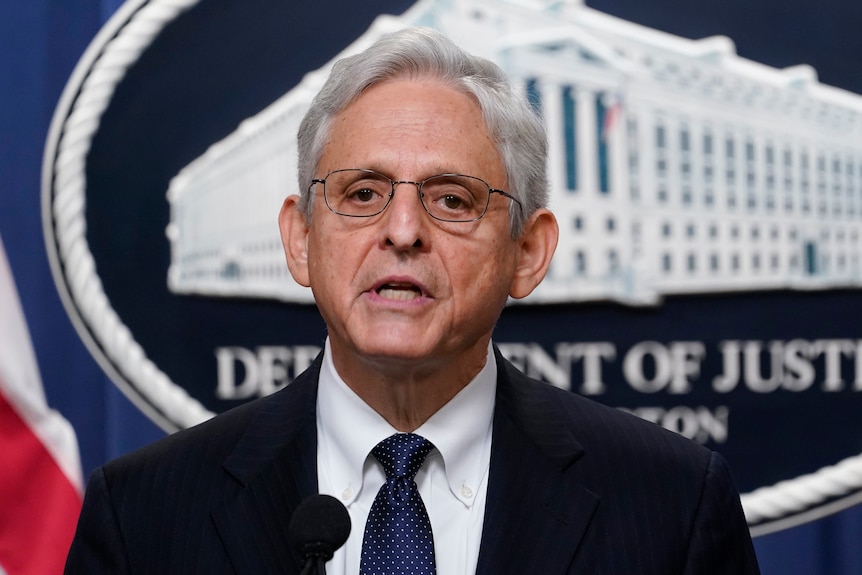 A serious-looking man with white hair and glasses speaks in front of the backdrop for US Justice Department press conferences.