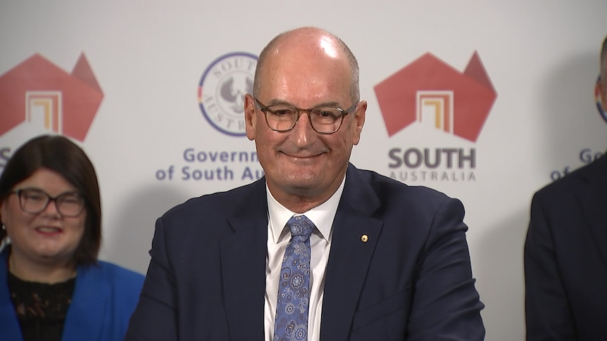 A man in a suit and glasses smiles while standing in front of a minister and a Government of South Australia banner