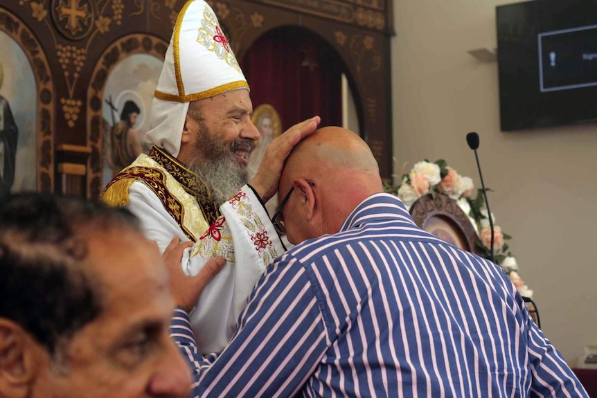The priest at the Coptic Orthodox Church in Taree, wearing white robes, places his hand on the head of a man in the church.