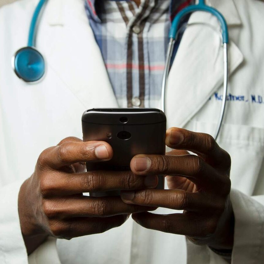 The torso of a person wearing a medical coat and stethoscope and holding a smartphone.