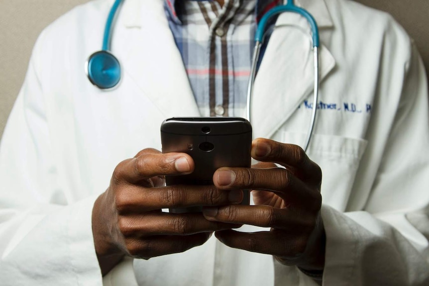The torso of a person wearing a medical coat and stethoscope and holding a smartphone.