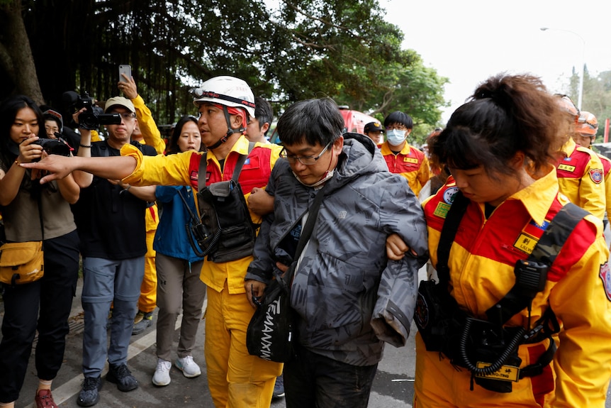 A man wearing a grey puffer jacket is supported by a two workers dressed in yellow jumpsuits on a busy street.