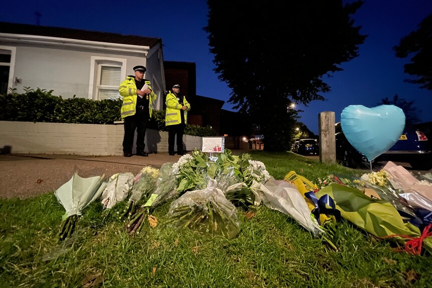 Two police in orange jackets are seen in the background, with floral tributes in the foreground