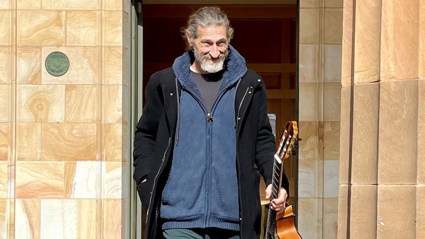 A man walks from a court building holding a guitar and smiling
