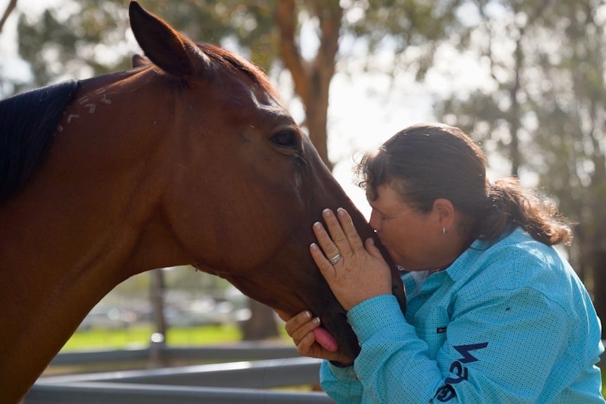 A woman wearing a blue shirt kisses the muzzle of a brown horse.