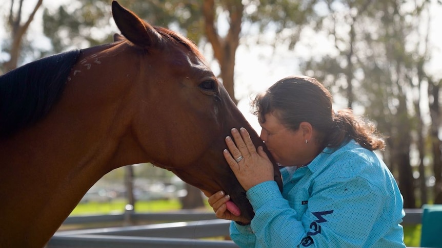 A woman wearing a blue shirt kisses the muzzle of a brown horse.
