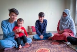 A woman in a veil sits on a colourful rug surrounded by two teen boys and a baby 