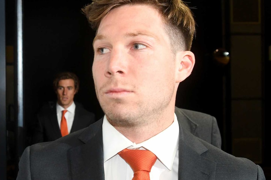 A male AFL player wearing a black suit, white shirt and orange ties looks to his right as he enters a building.