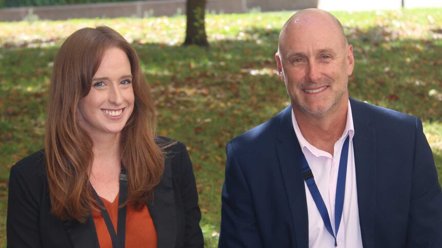 An auburn-haired woman in a suit sits beside a bald-headed man in a suit.