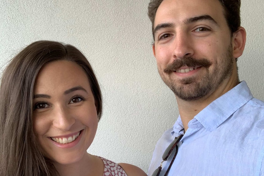 A brunette woman stands next to a man with a moustache. They're both smiling.