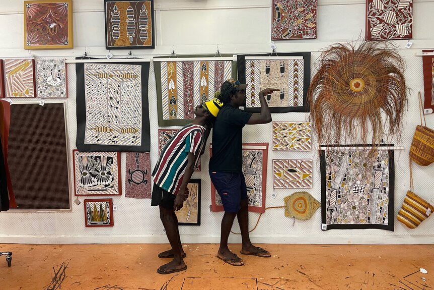 Two young Aboriginal men strike funny poses. They are standing in front of a wall covered in art. 