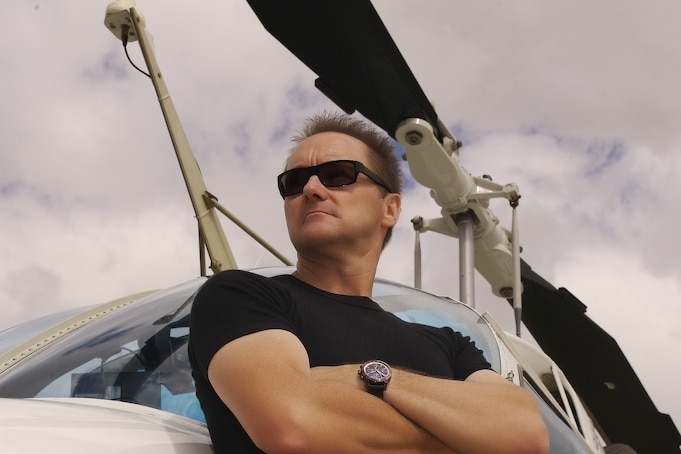 A man in sunglasses and a muscle shirt leans against a
            helicopter.