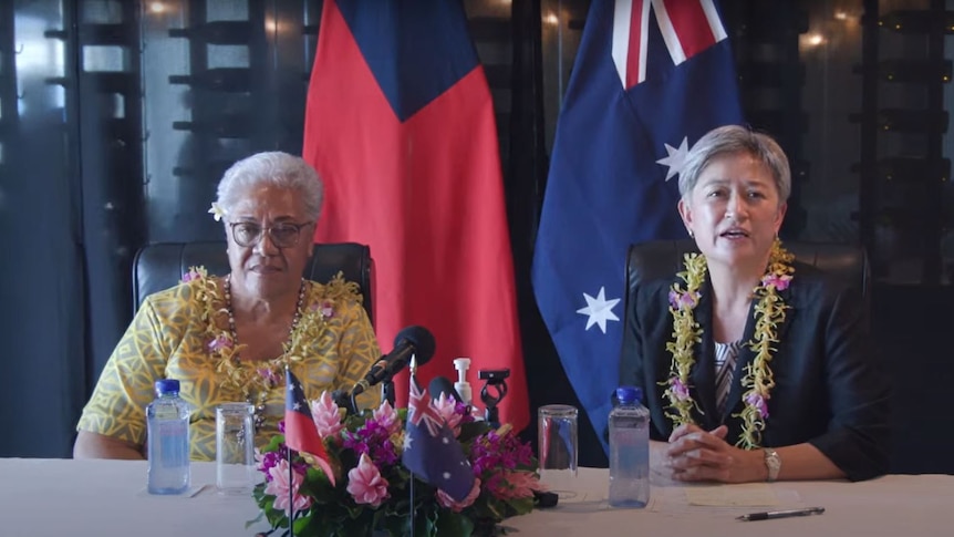 Two women sit at a table in front of the flags of Samoa and Australia