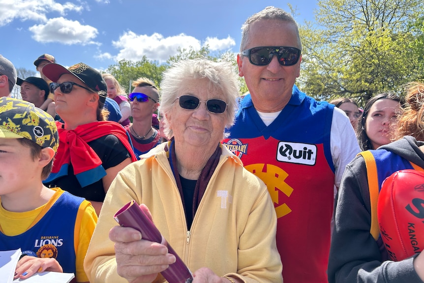 A woman with white hair and sunglasses wears a yellow jumper and Lions scarf while her son stands behind in a Fitzroy guernsey.