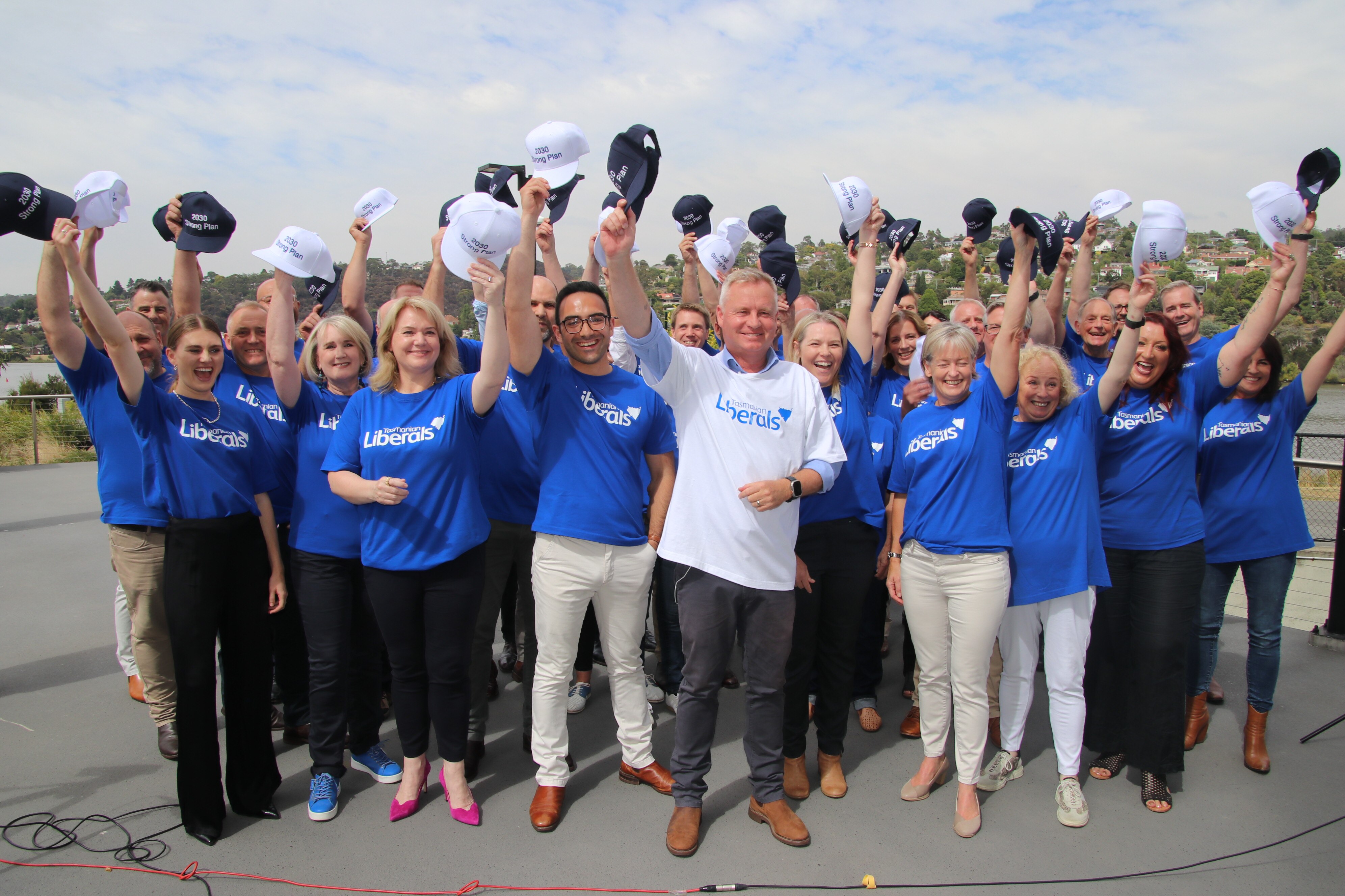 A man wearing a light blue liberal shirt stands at the front of a group of people who are all holding their caps in the air.