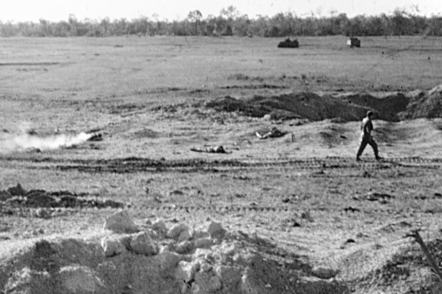 There is a man standing on a baron battlefield with tanks in the background.
