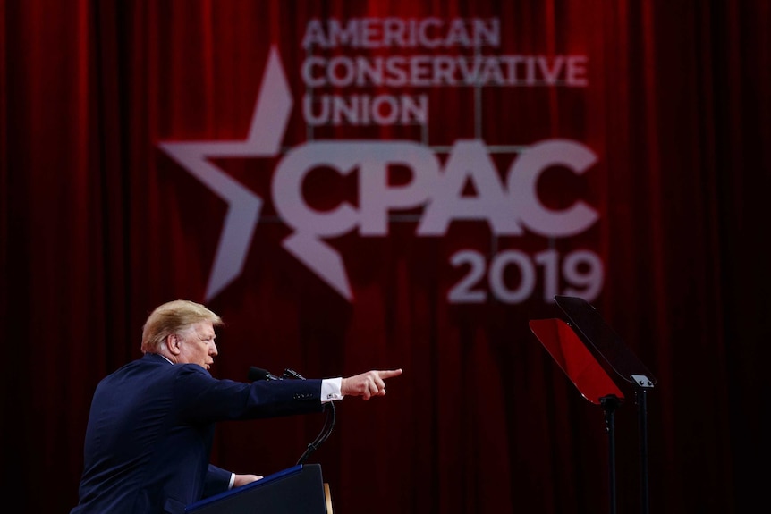 Donald trump stands on stage behind a podium speaking into a microphone and pointing forward