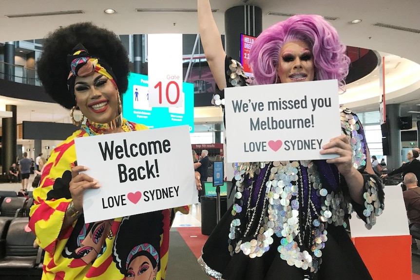 Two drag queens pose in Sydney Airport with signs that read 'Welcome back!'