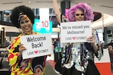 Two drag queens pose in Sydney Airport with signs that read 'Welcome back!'