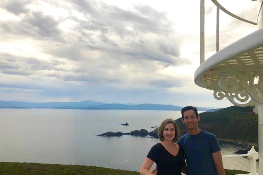 A couple with their arms around each other, standing on a high vantage point overlooking water and mountain ranges.