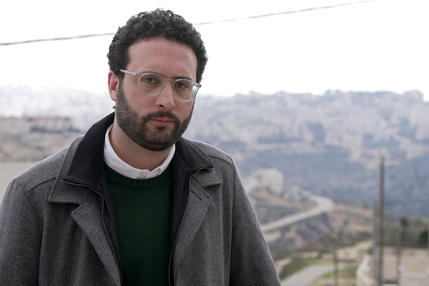 A man wearing glasses stands outside with a serious expression. Behind him are low mountains and a road.