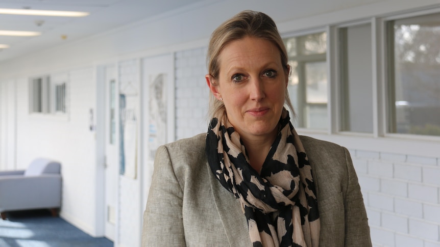 A blonde woman in a scarf stands in a hallway.