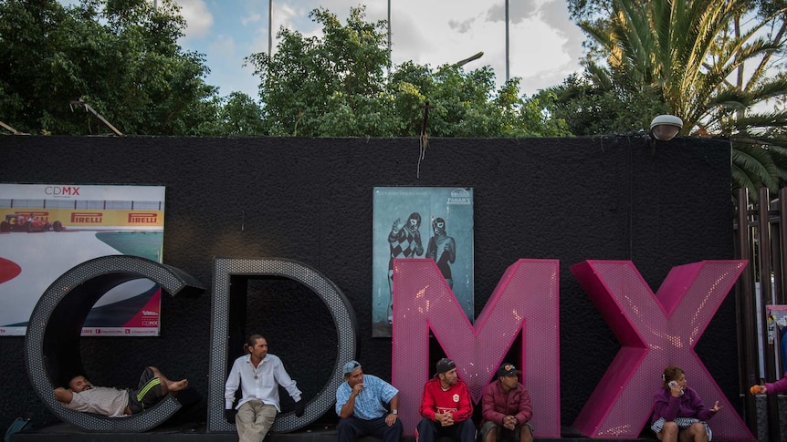 Central American migrants rest and phone home while sitting in big letters spelling CDMX